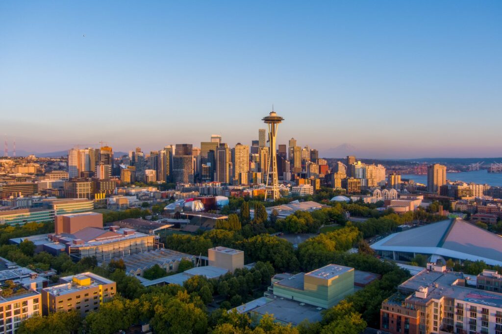 Depicts a wide, elevated view of the Seattle skyline at sunset, showcasing the iconic Space Needle rising prominently above surrounding skyscrapers and a lush green park in the foreground under a clear sky.