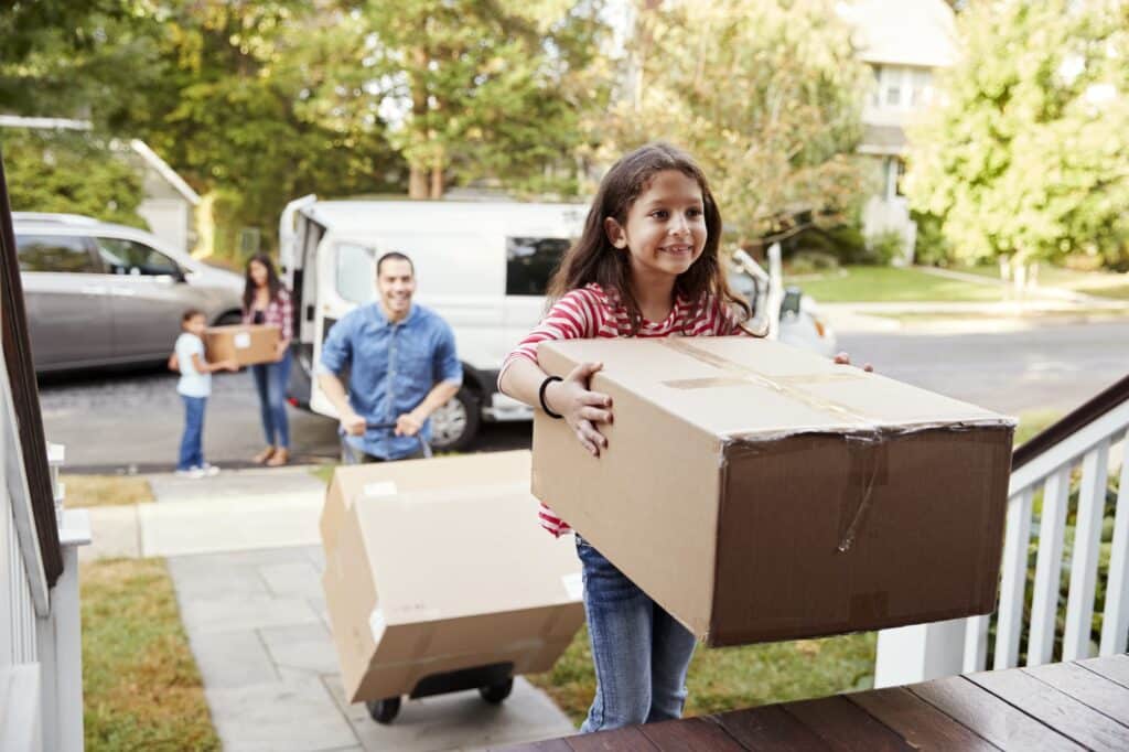 A smiling young girl carries a large cardboard box up the front porch steps while her family unloads a moving van in the background.