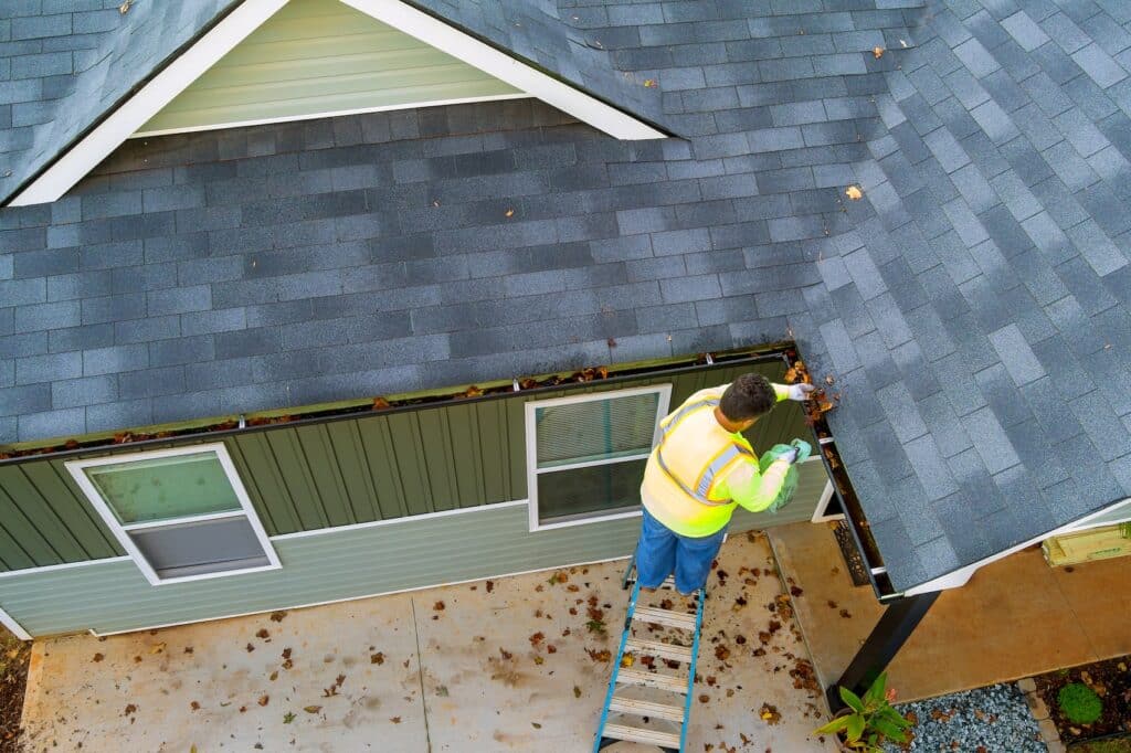 An overhead view of a worker wearing a high-visibility vest and standing on a ladder while cleaning fallen leaves out of the rain gutters of a residential house.