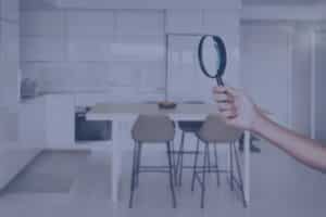 A hand holds a magnifying glass against a blurred background of a modern kitchen and dining area.