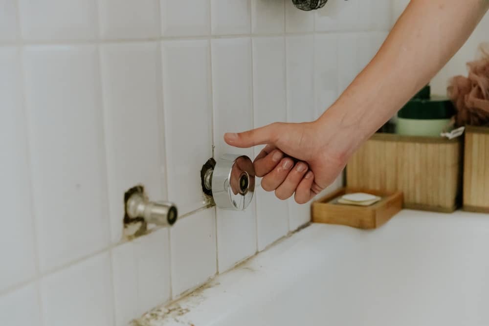 A hand reaches to turn a chrome fixture on a white tiled shower wall with visible mold and grime around the bathtub edge.